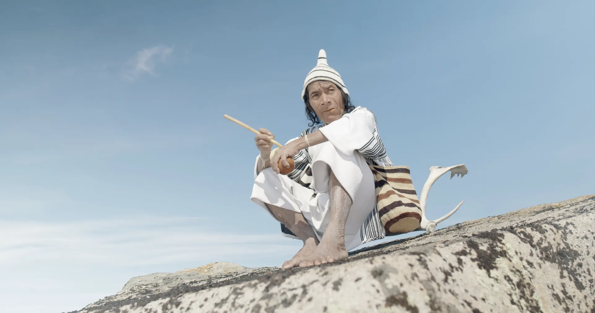 An elder in white clothing and a conical hat crouching on a rock beside a reindeer antler under a blue sky